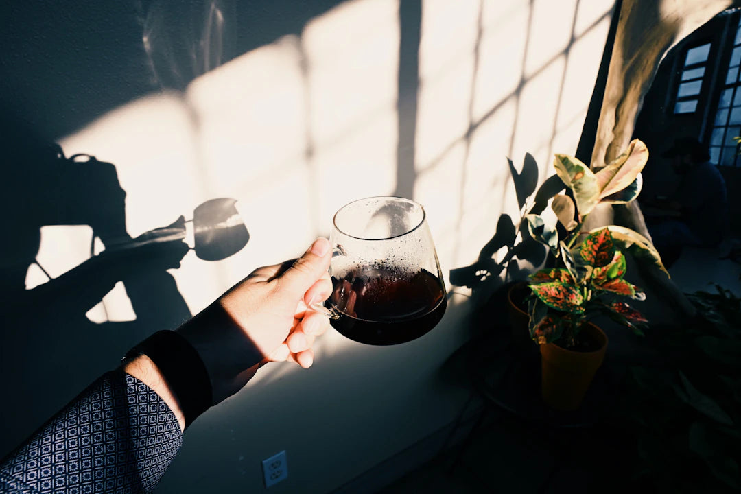Hand holding a glass of dark liquid with sunlight shadows.