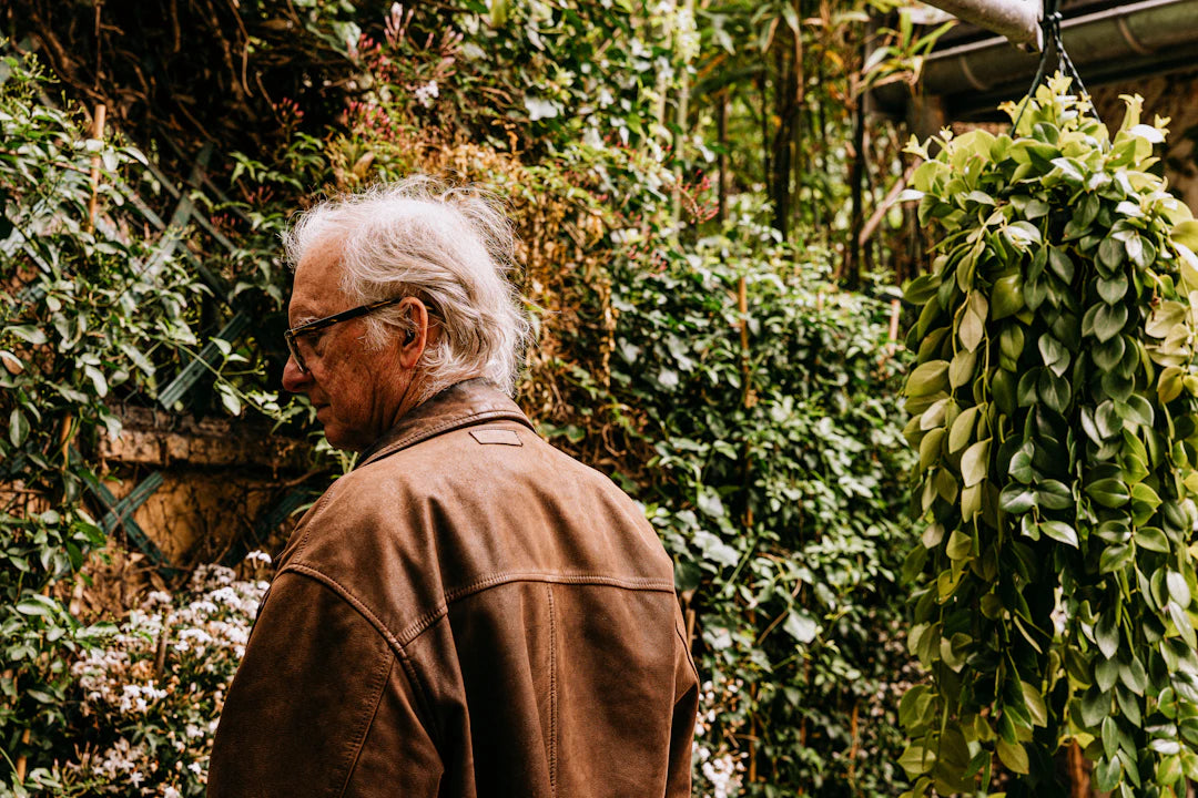 Elderly man in brown jacket surrounded by lush greenery